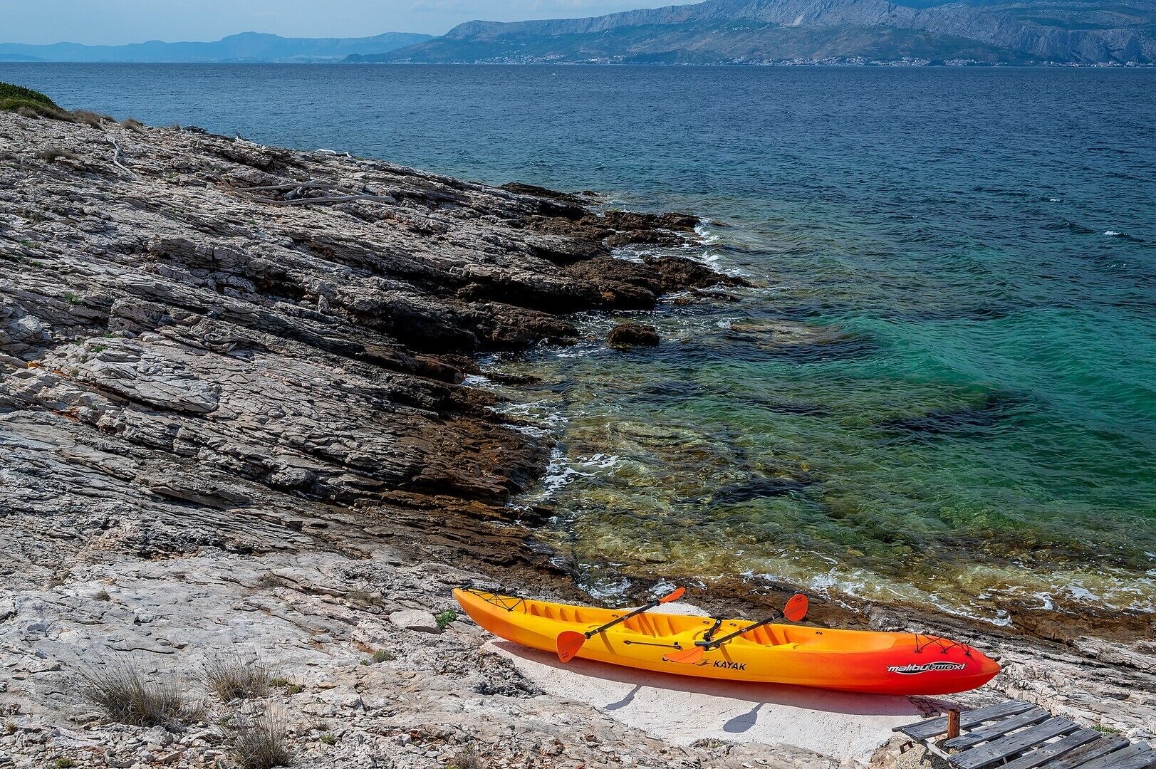 Un kayak en una costa rocosa con aguas claras y montañas al fondo.