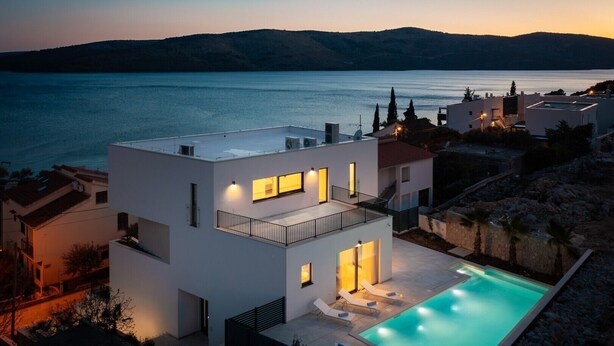 Et moderne hus med to etager og swimmingpool, beliggende nær vand med bjerge i baggrunden.