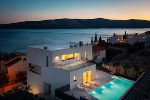 A modern two-story house with a swimming pool, located near a body of water with mountains in the background. - NEW ! Villa Brilliance, heated pool 50m2 sea view, big group 100 m from sea, bbq (Seget Vranjica)