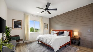 A bedroom with a bed, a ceiling fan, a window with curtains, a bedside table with a lamp, and a plant.