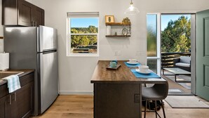 A modern kitchen with a stainless steel refrigerator, wooden cabinets, a central island with seating, and a view of a garden through a window.