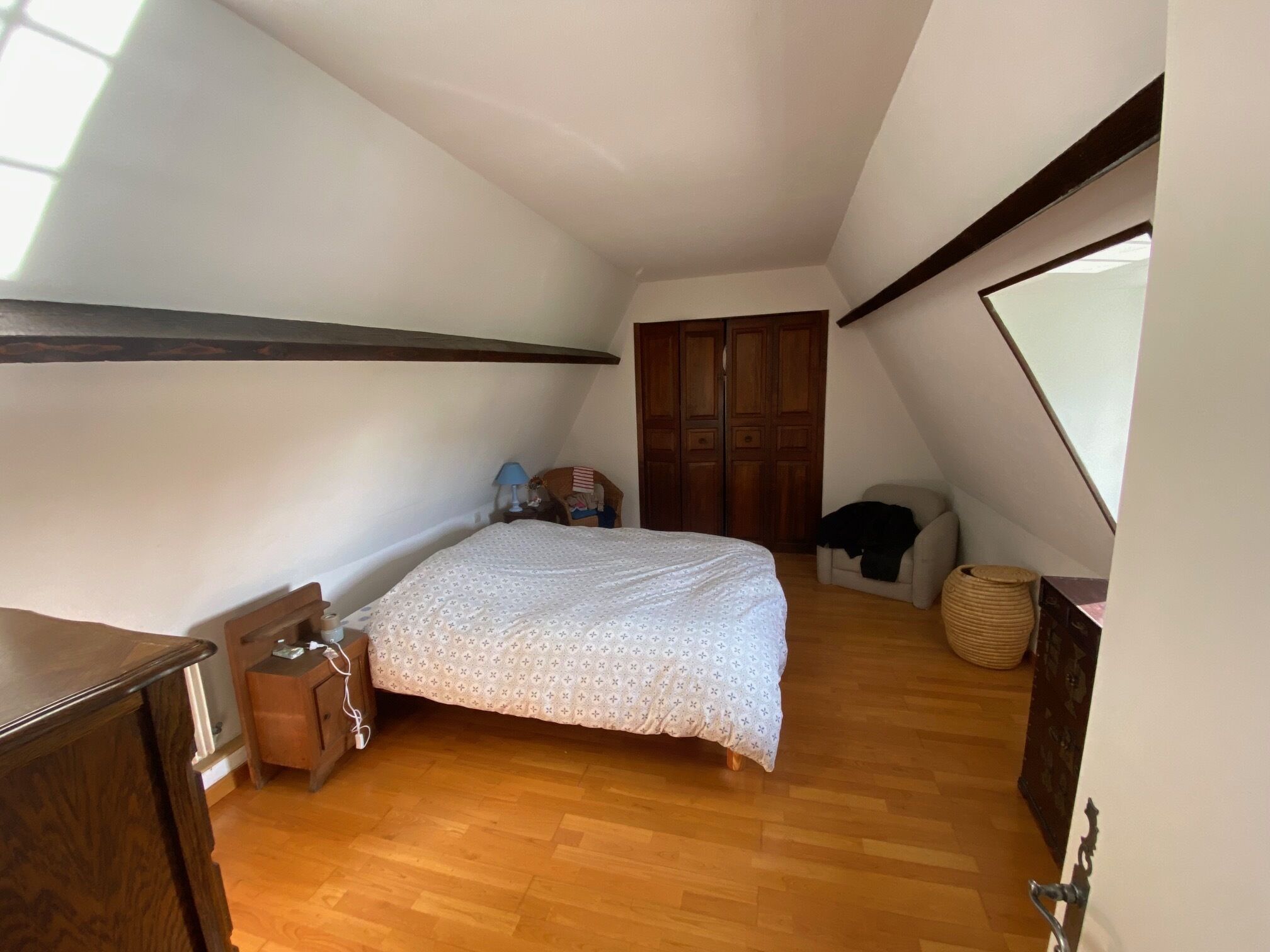 A bedroom with a wooden floor, a bed with a patterned bedspread, a nightstand with a lamp, a wooden wardrobe, and a window with a view of the outside.