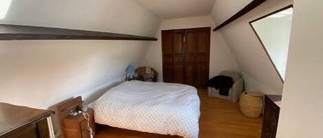 A bedroom with a wooden floor, a bed with a patterned bedspread, a nightstand with a lamp, a wooden wardrobe, and a window with a view of the outside.