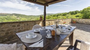 A wooden table set for dining with white dishes and cups, surrounded by stone walls and a view of green hills. - Apartment Marcigliana Front Pool Radicondoli (Radicondoli)