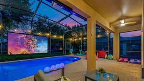 A swimming pool area with a large screen displaying a video game, surrounded by a covered patio with seating and a table.