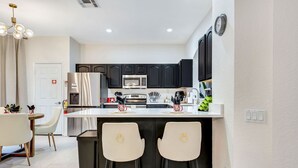A modern kitchen with dark cabinets, a white countertop, and a stainless steel refrigerator. There are two bar stools with gold rings and a small island with a coffee machine.