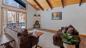 A living room with a fireplace, wooden ceiling beams, a sofa, a table, and a view through large windows.