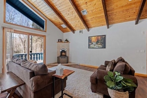 A living room with a fireplace, wooden ceiling beams, a sofa, a table, and a view through large windows.
