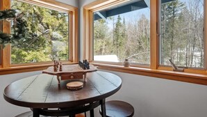 A round wooden table with chess pieces and a vase, set in a room with large windows overlooking a forest.