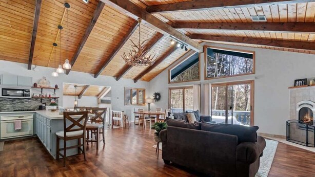 A spacious living area with a fireplace, wooden beams, and a large window overlooking a forest.