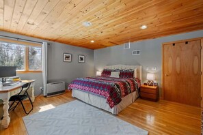 Ein Schlafzimmer mit Holzboden, einem Bett mit gemustertem Überwurf, einem Schreibtisch mit Computer und einem Fenster mit Blick auf Bäume.