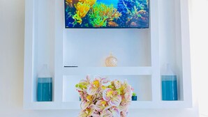 Una sala de estar moderna con un televisor de pantalla plana montado en una pared blanca, un jarrón decorativo con flores sobre una mesa de vidrio, y una bandeja con tazas y una tetera.