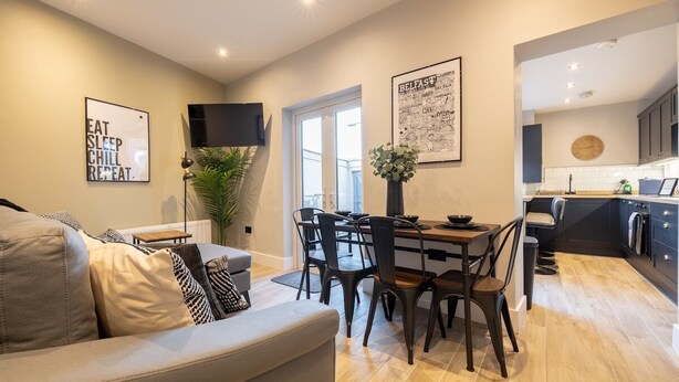 Ein modernes Wohnzimmer mit einer Couch, einem Esstisch und Stühlen. Es gibt eine Küchenzeile mit Schränken und einer Uhr an der Wand.