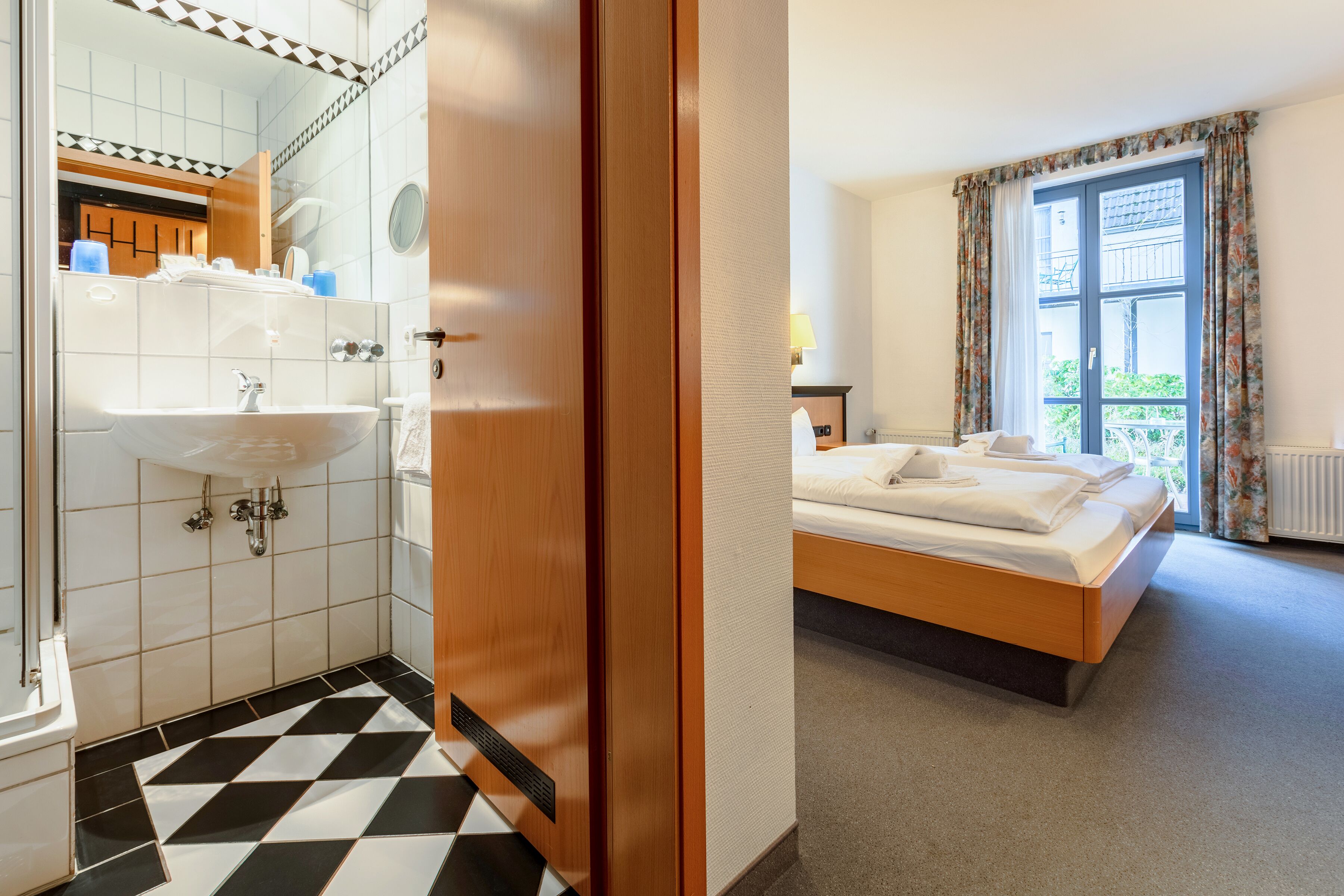 A hotel room with a wooden bed, a bathroom with a sink and mirror, and a checkered floor.