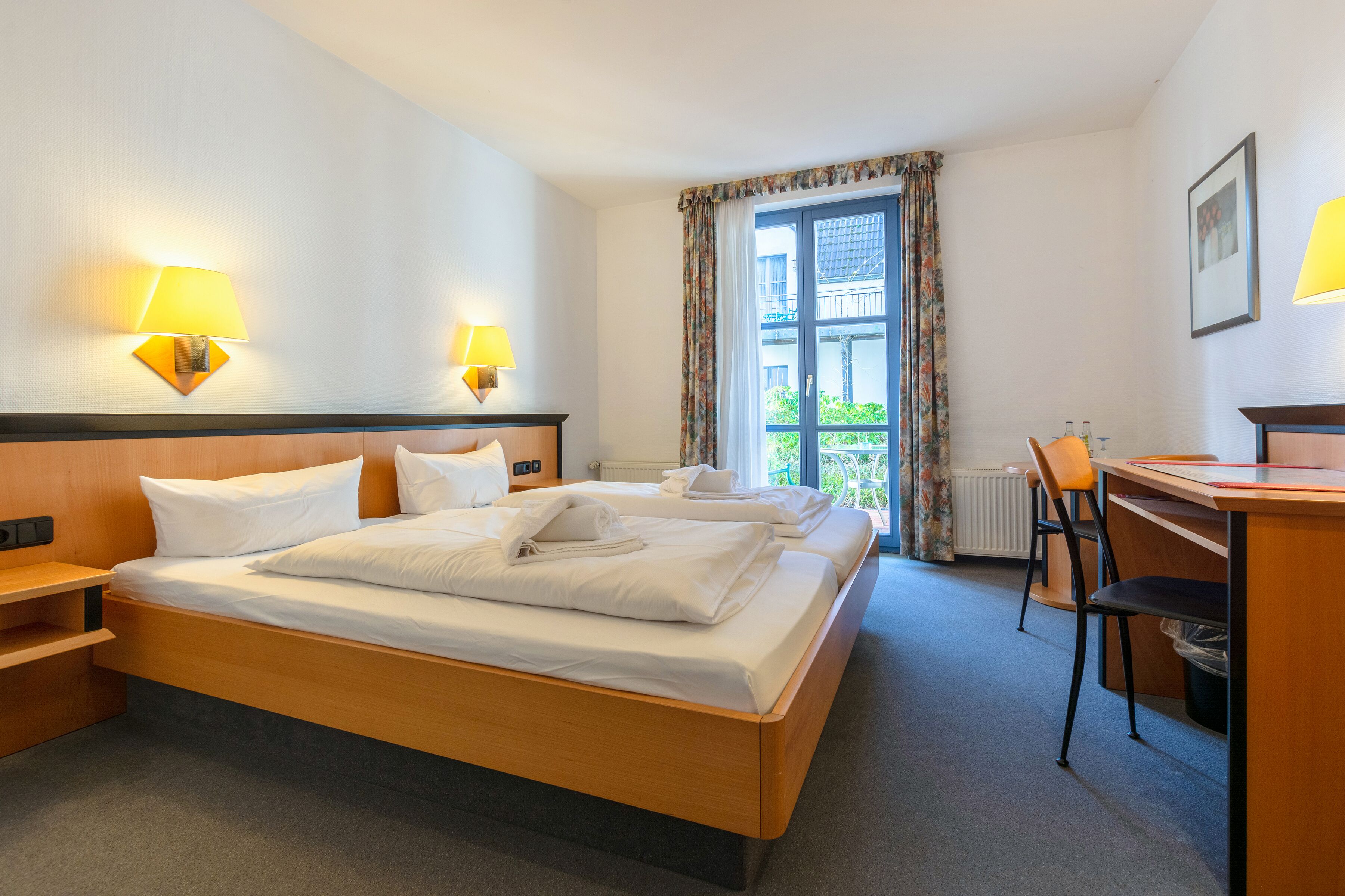 Habitación de hotel con cama doble, dos lámparas de noche, un escritorio con silla y vistas al exterior a través de una ventana con cortinas.