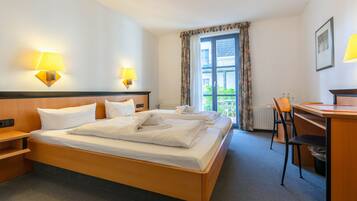 A hotel room with a double bed, two bedside lamps, a desk with a chair, and a view of the outdoors through a window with curtains.