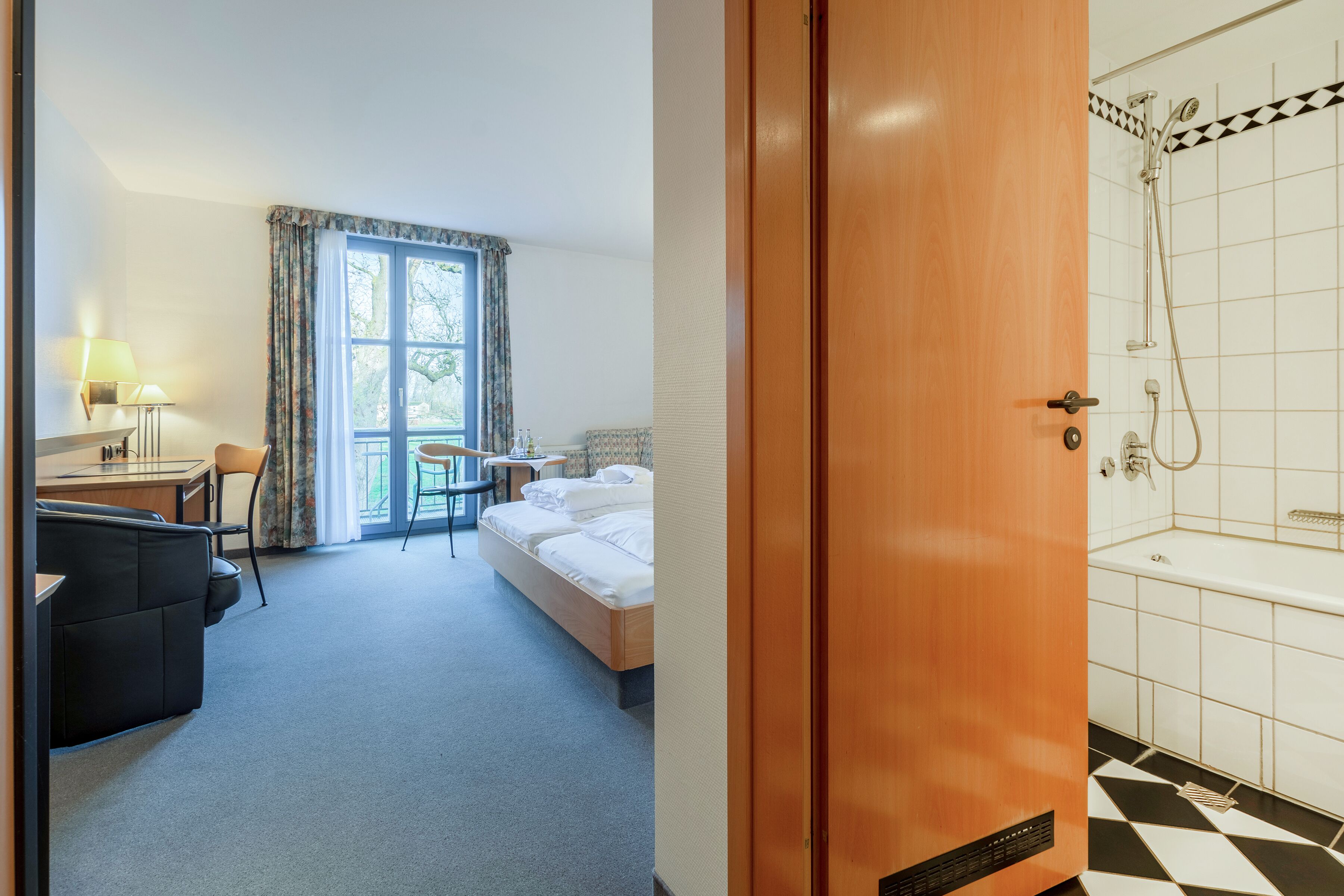 Hotellihuone, jossa on sänky, työpöytä tuolilla, ikkuna verhoilla sekä kylpyhuone, jossa on suihku ja kylpyamme.