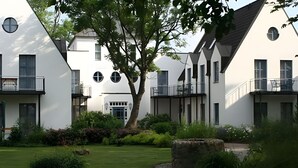 A modern building with a symmetrical facade, multiple balconies, and a well-maintained garden with a central tree and stone feature. - Hotel Rügenschmiede vis a vis Hiddensee (Schaprode)