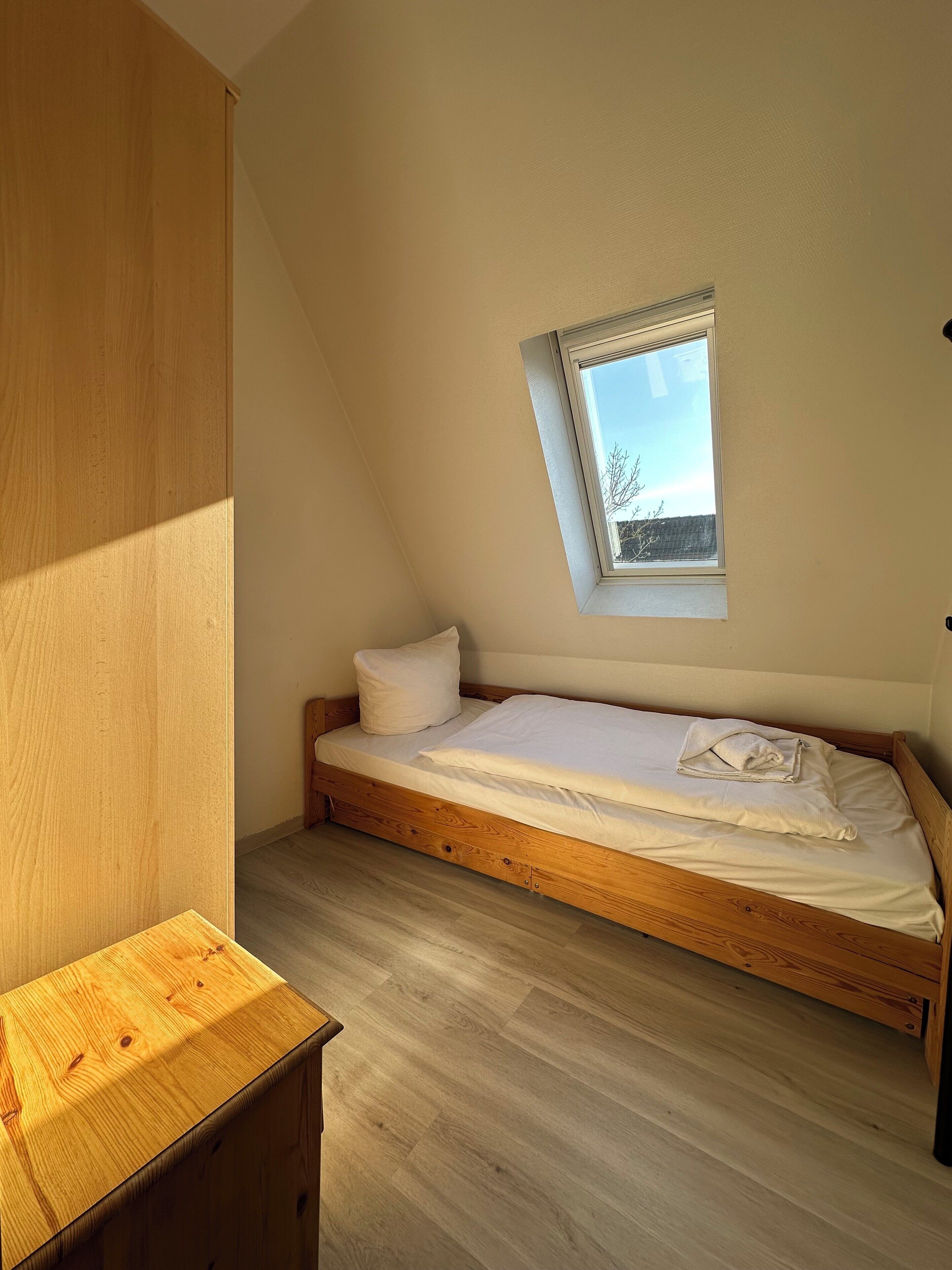A small, single-bed room with a sloped ceiling and a skylight, wooden flooring, and a simple bedside table.
