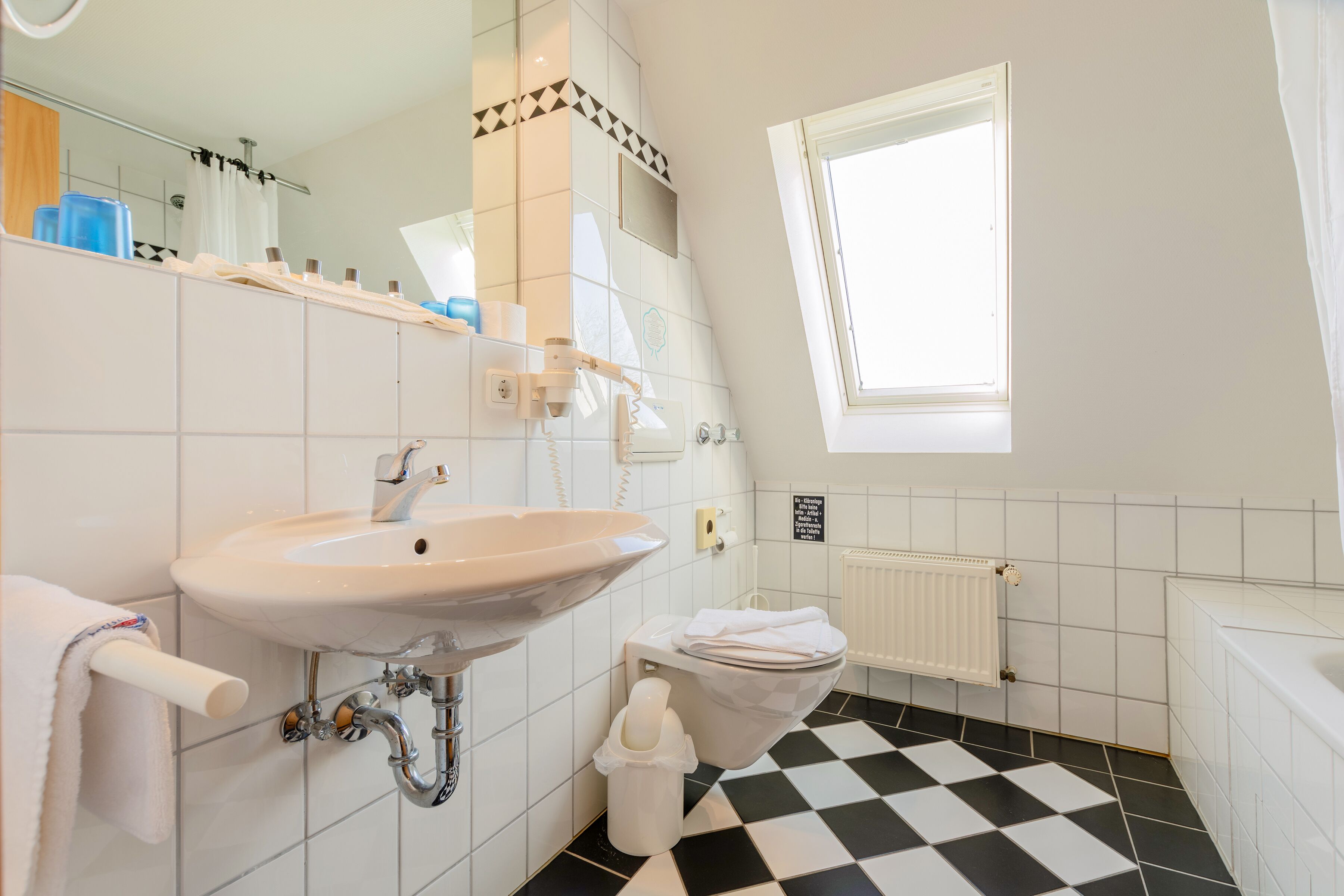 Ein Badezimmer mit weißem Waschbecken, Toilette und Badewanne. Ein Fenster sorgt für natürliches Licht und der Boden ist kariert.