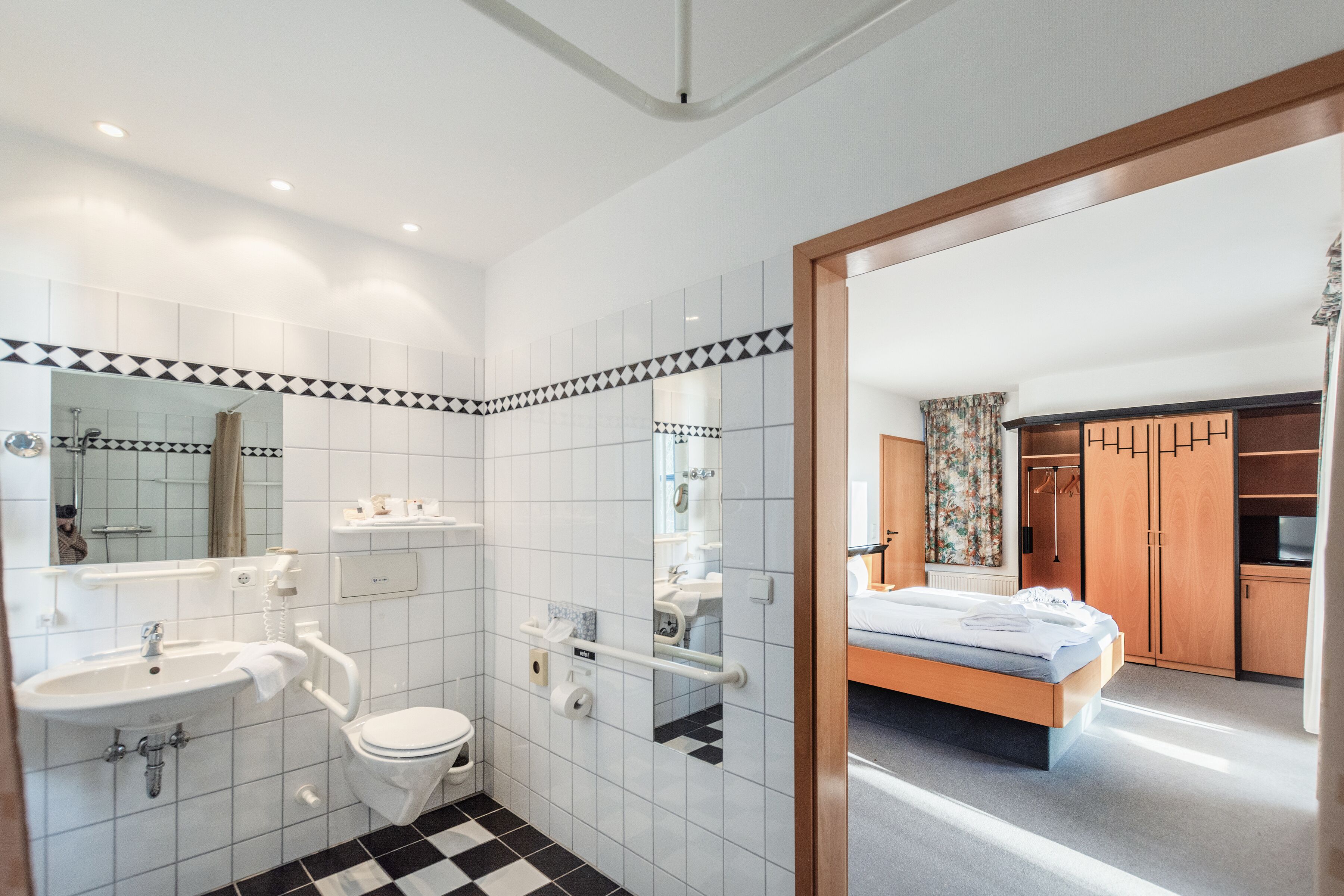 Une salle de bain moderne avec un lavabo, des toilettes et un miroir. Le sol est carrelé et une porte en bois donne sur une chambre à coucher équipée d’un lit et d’une armoire.