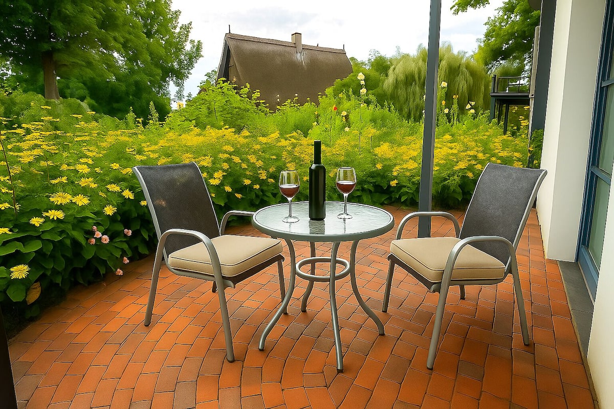 A patio with a round table and two chairs, surrounded by a garden with yellow flowers and a building in the background.