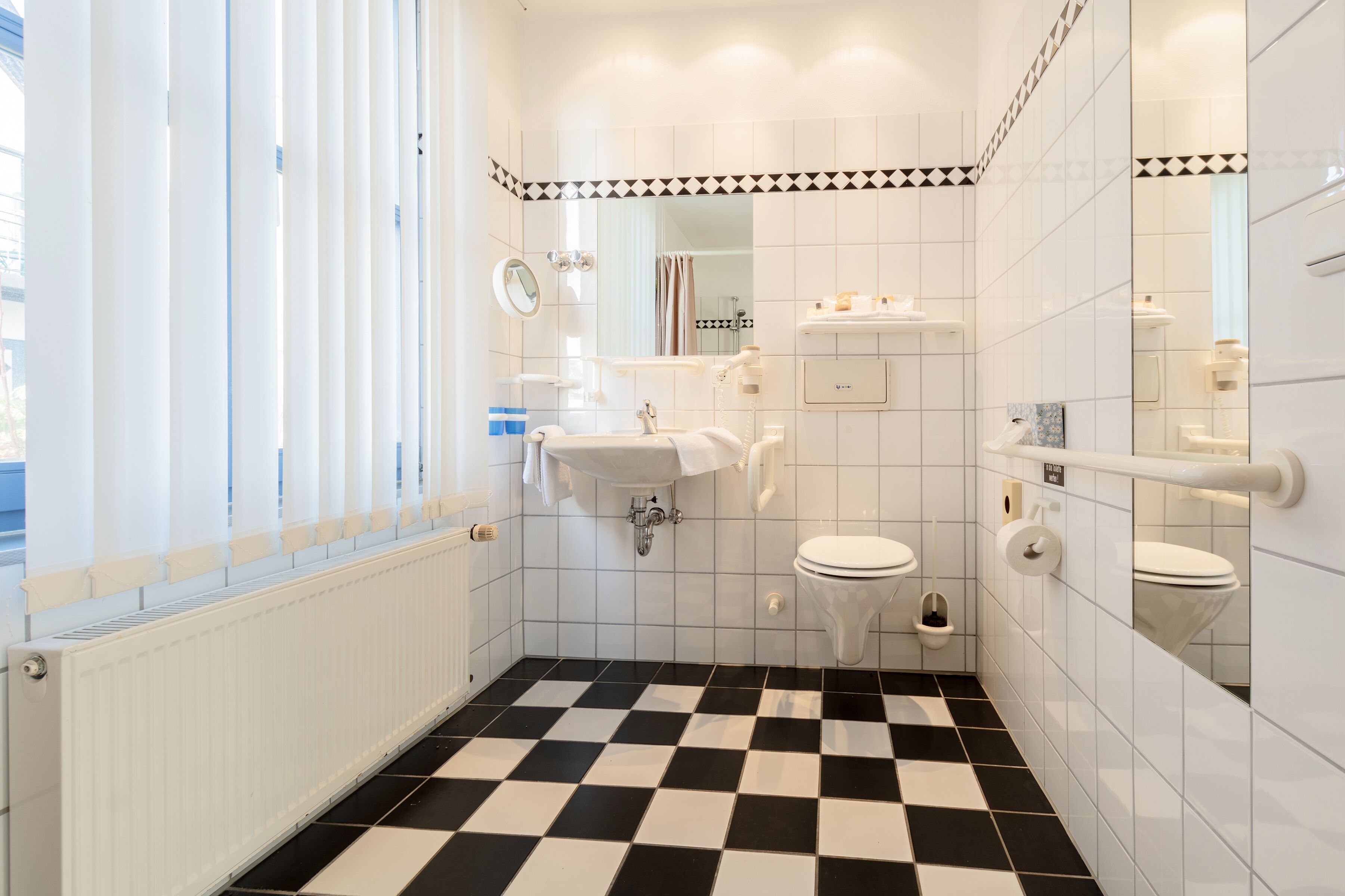 A bathroom with a black and white checkered floor, white tiles, a sink, a toilet, a mirror, and a window with blinds.