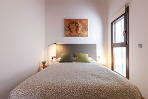 A bedroom with a patterned bedspread, a headboard, a bedside table with a lamp, a wall-mounted artwork, and a window. - Iconic Old Town - Unik Vacation (Tarragona)
