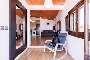 A room with a wooden floor, a rocking chair, a sofa, a washing machine, and a kitchen area.