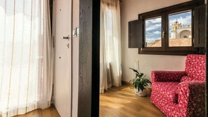 Um quarto com piso de madeira, um sofá floral vermelho, uma janela com vista para um edifício e um vaso de planta.