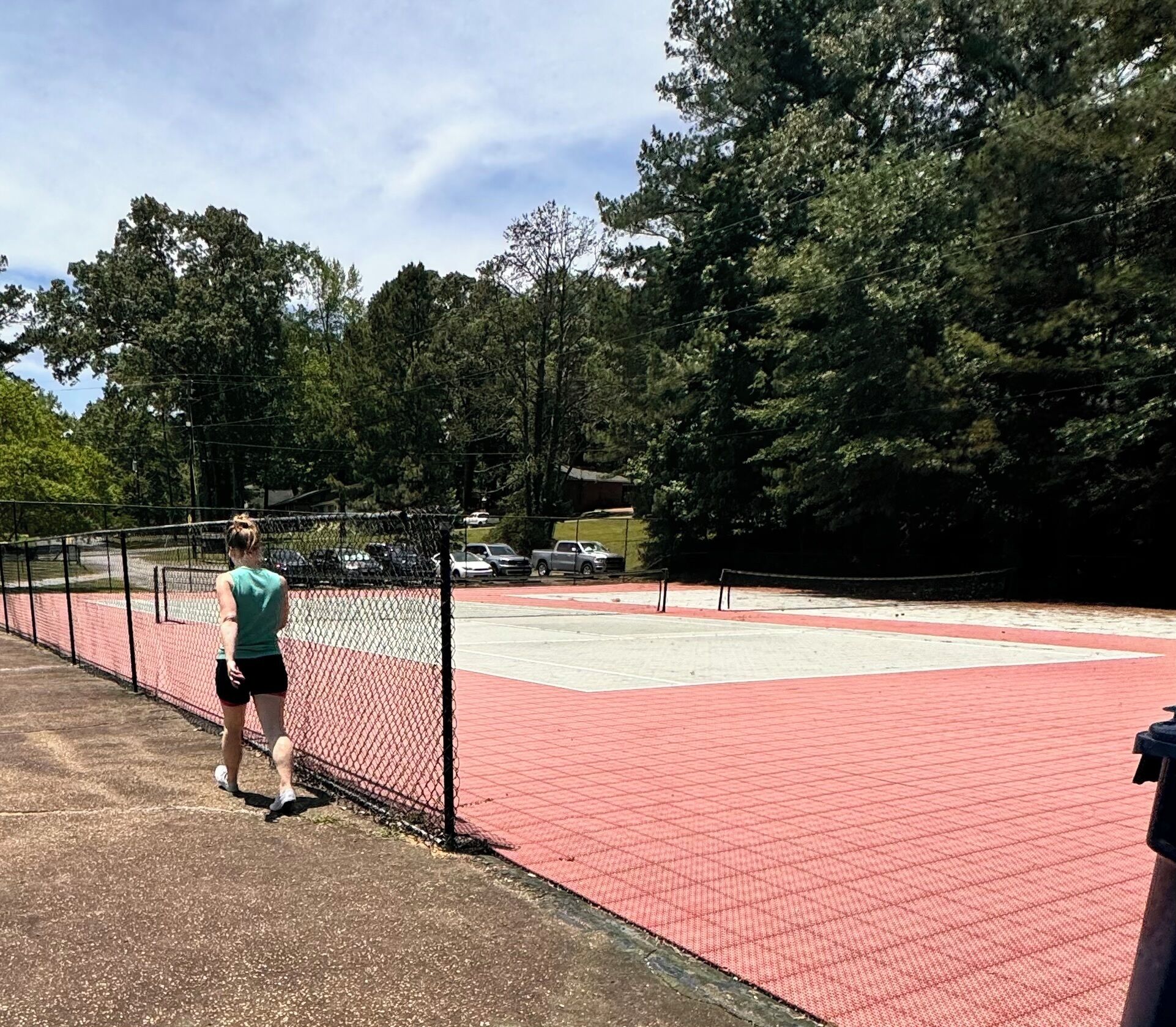 Una cancha de tenis con cerca de malla, superficie roja y árboles al fondo.