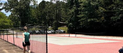 Una cancha de tenis con cerca de malla, superficie roja y árboles al fondo.