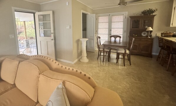 Una sala de estar con un sofá color beige, una mesa de comedor con sillas, una zona de cocina con un armario y un reloj, y una puerta que da al exterior.