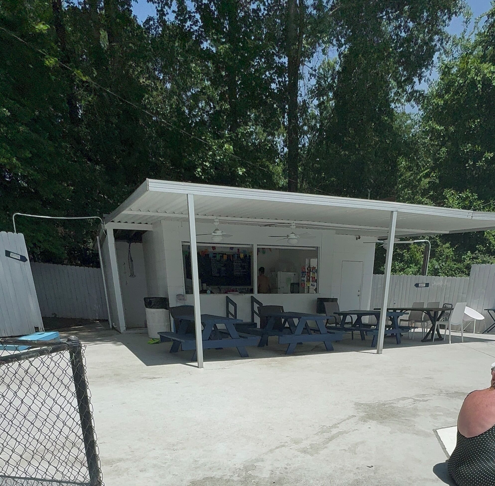 A covered outdoor seating area with tables and chairs, a white building with a window, and a chain-link fence.