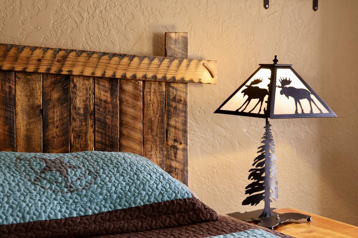 Een bed met een blauw-bruine quilt, een houten hoofdbord en een wandlamp met silhouet van een eland.