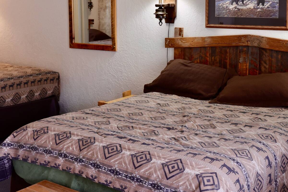 Een slaapkamer met een bed, een houten hoofdbord, een nachtkastje, een spiegel, een wandlamp en een ingelijste foto.