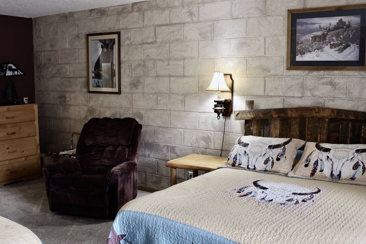 Een kamer met een stenen muur, een houten hoofdbord, een bed met bedkussens met een patroon, een houten stoel, een klein tafeltje en een lamp.