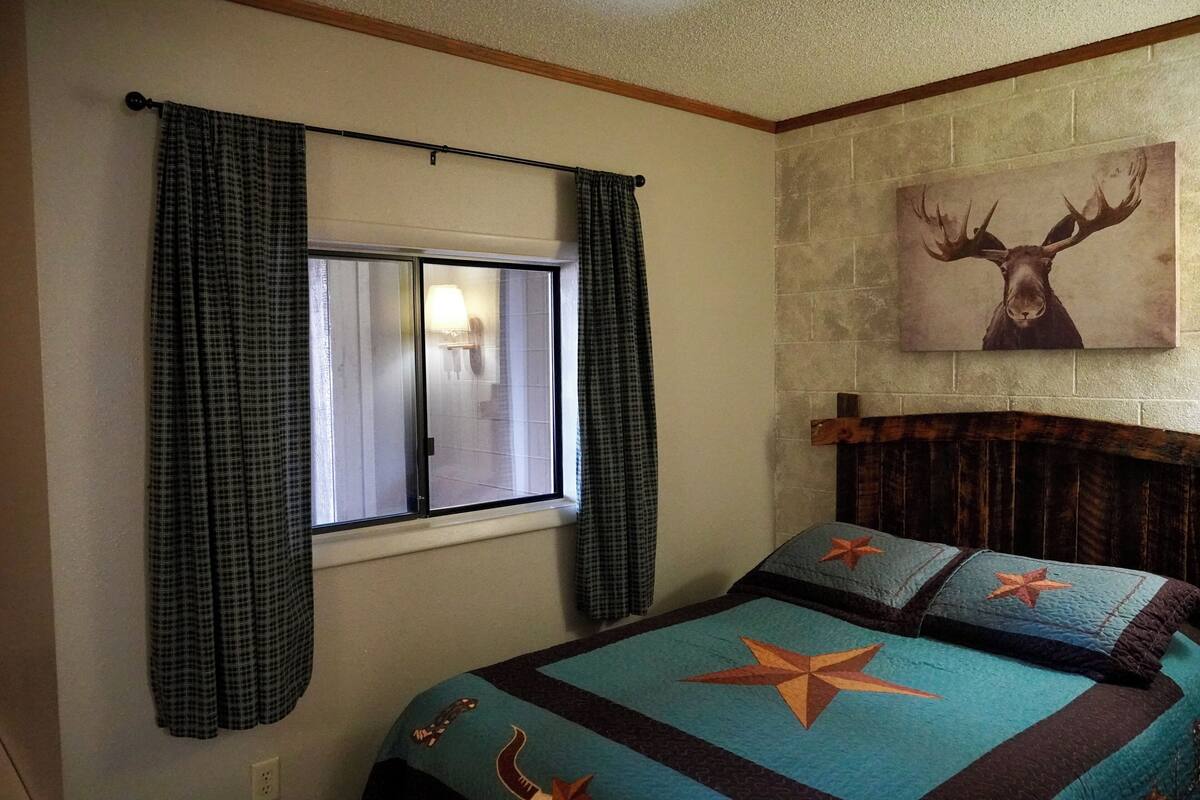 Een slaapkamer met een bed, geruite gordijnen, een raam en een aan de muur hangend hert schilderij.