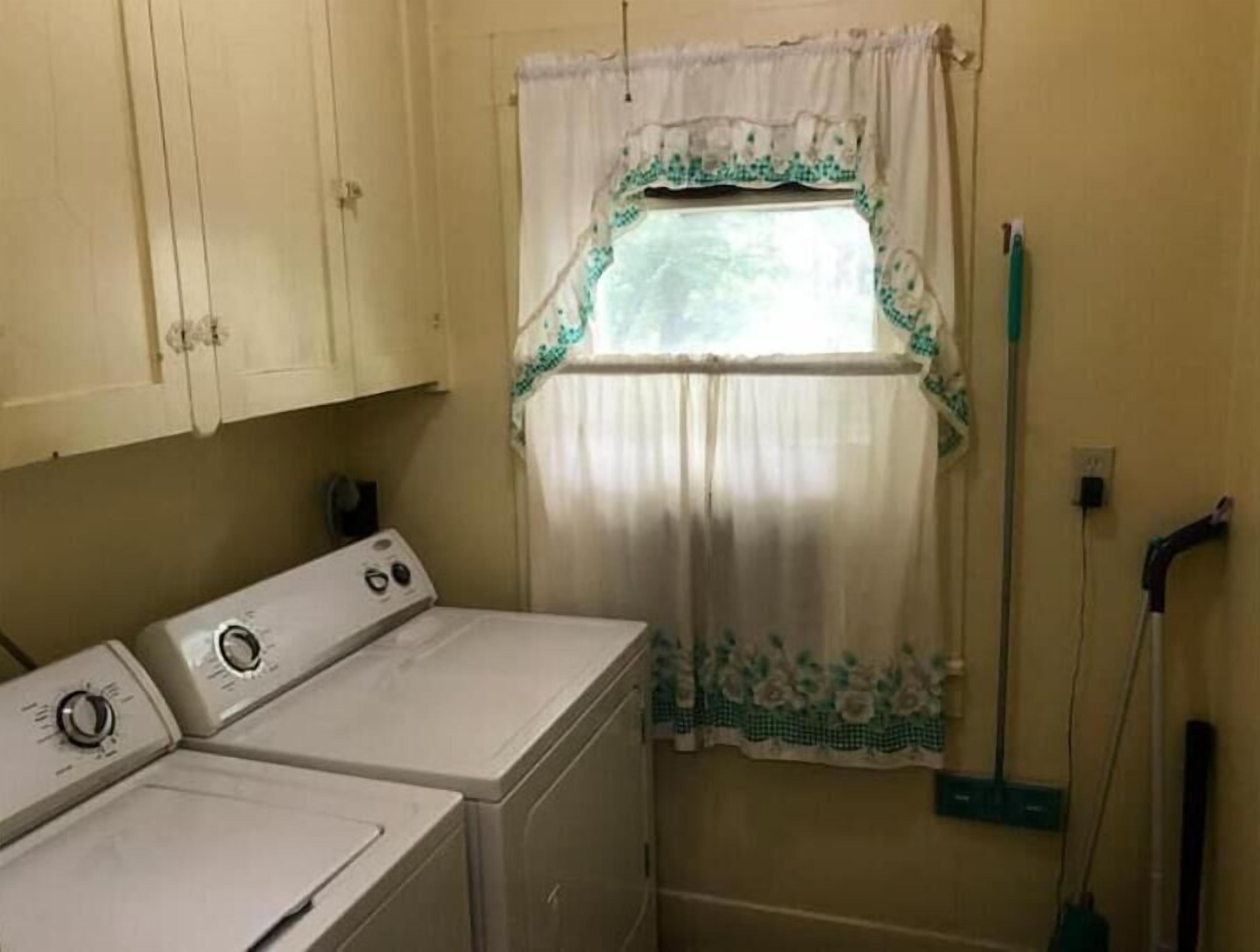 A laundry room with two stacked washers and dryers, a window with floral curtains, and a mop leaning against the wall.
