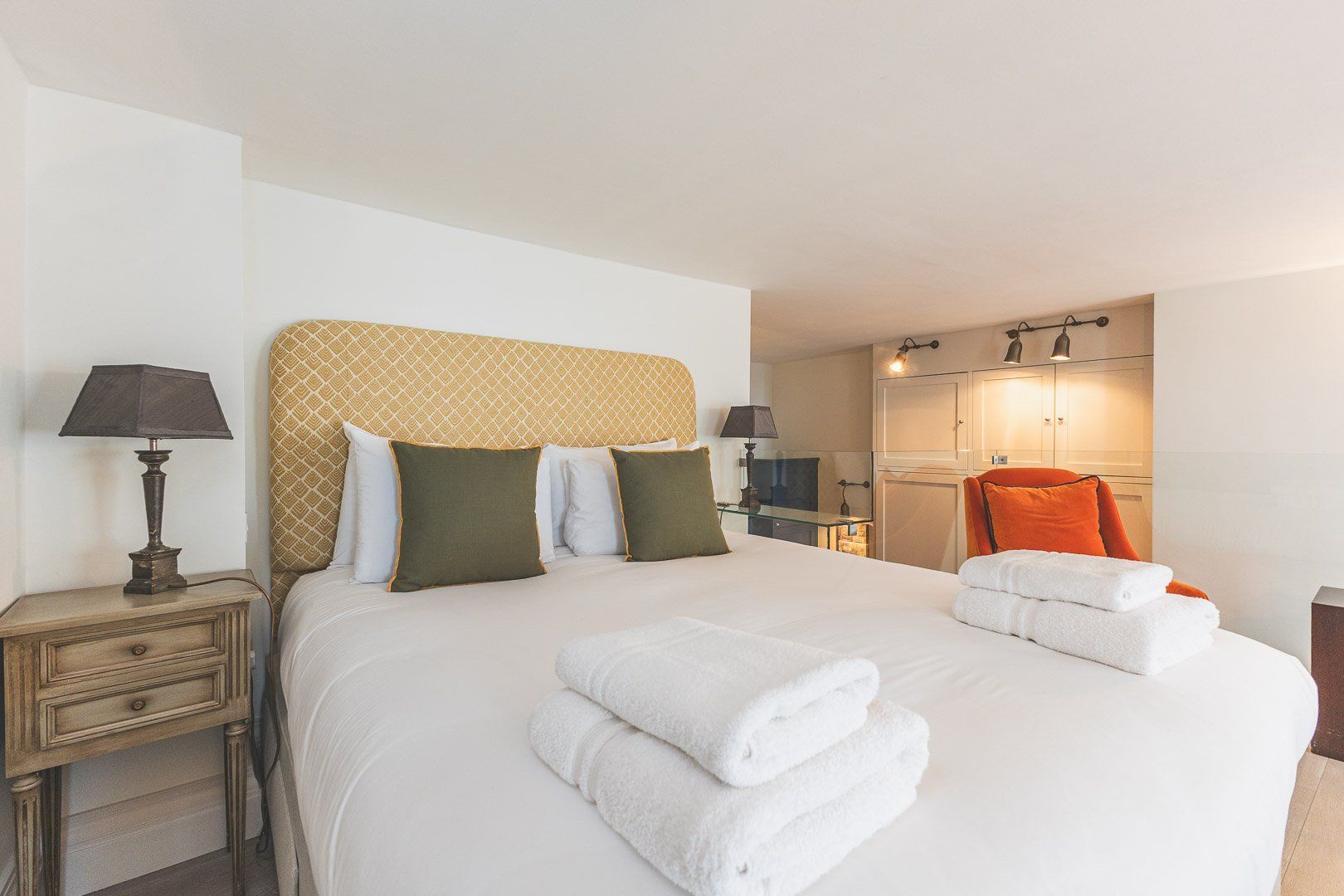 A hotel room with a large bed, two bedside tables, a nightstand, a wardrobe, and a bathroom with a mirror and towel rack.