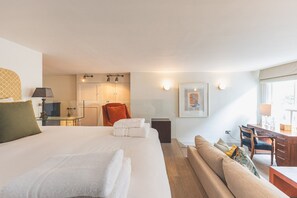 Habitación de hotel con una cama grande, un sofá, un escritorio y una silla. Se observa una peque ña kitchenette al fondo.