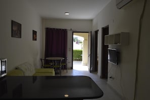 A room with a dining table and chairs, a sofa, a flat-screen TV mounted on the wall, and a door leading outside. - Nice 2-bed Apartment (Makadi Bay)