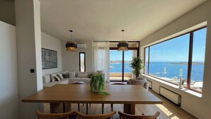 Een ruime eethoek met een grote houten tafel en vier stoelen, een bank en uitzicht op zee door grote ramen.