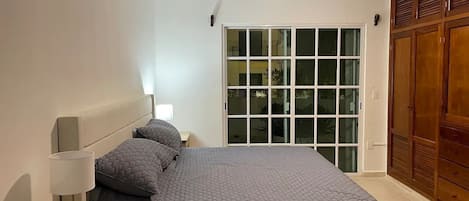 Un dormitorio con cama, mesita de noche, lámpara, ventilador de techo y puerta corrediza de vidrio que da a un balcón.