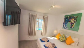 A hotel room with a bed, a TV, a painting, and a window with curtains. - Casa Bill | Firepit Nights by Villas Now (Playa Blanca)