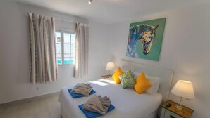 A hotel room with a bed, two bedside lamps, a window with curtains, and a colorful painting on the wall. - Casa Bill - Firepit Nights by Villas Now (Playa Blanca)
