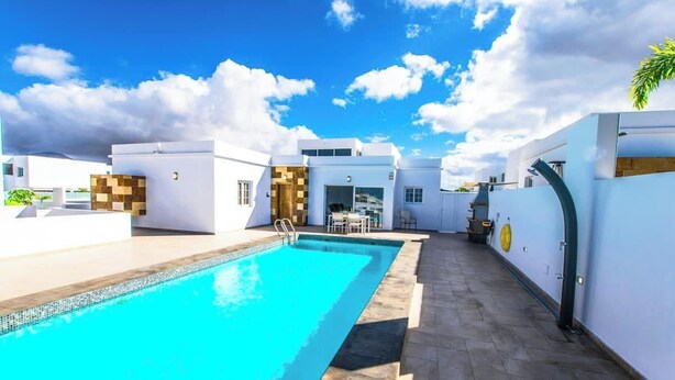 A modern pool area with a clear blue pool, white walls, and a covered seating area. - Casa Bill - Firepit Nights by Villas Now (Playa Blanca)