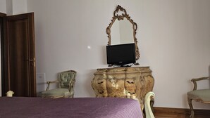 Une pièce comprenant un lit, un téléviseur posé sur un meuble en bois, deux fauteuils rembourrés et une porte.