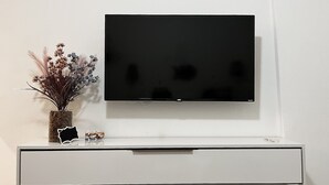 Un televisor de pantalla plana montado en una pared blanca sobre un mueble blanco con un jarrón de flores secas y un par de gafas.