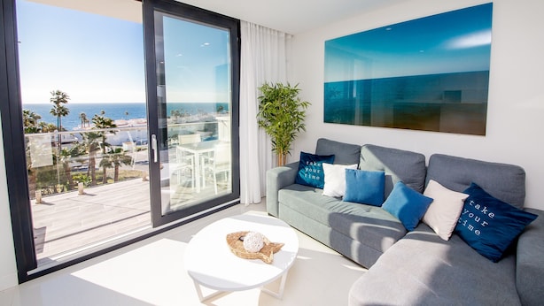 A modern living room with a grey sofa, white coffee table, and a large window offering a view of the sea and palm trees.